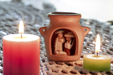 Velas decorativas de color rosa y verde con llamas encendidas sobre una alfombra tejida a crochet blanco junto a un pesebre de origen latinoamericano. Detalle de Navidad.