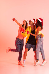 Obraz premium happy girl taking selfie with multicultural friends in santa hats on pink
