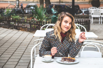 Beautiful young Caucasian having a rest with cappuccino and a piece of cake on the street table of a small cafe on an autumn day.