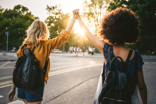 Couple of young women from the back, holding hands with arms raised and they walk in the street at sunset - Two millennials are happy