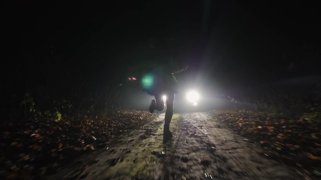 A frightened girl quickly runs away from a car following her along a country road at night. Silhouette of a running girl in the headlights of a car.