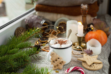 Fototapeta na wymiar Cozy autumn or winter still life on a windowsill with cocoa or coffee with marshmallows, gingerbread, spruce branches, dried citruses, pumpkin, Christmas candies and candles on a knitted background.