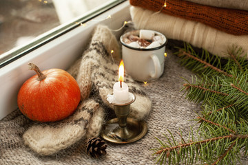 Candle in a bronze or metal candlestick burns out on the windowsill. Cozy still life on a gray background with knitted mittens, spruce branches, pine cone, pumpkin, cup with a hot drink and lights.