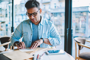 Mature contemplative hindu entrepreneur in glasses for eye protection drawing up piece of small paper sitting in coffee shop.Handsome skilled pensive male architect working indoors with tools