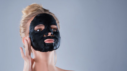 girl with naked shoulders posing at grey background with facial mask black, skin care concept, beauty photo, skin treatment, hydrating skin mask, smiling.