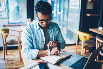 Fototapeta premium Mature good looking male entrepreneur dressed in casual denim clothes configuring smartwatch while waiting for meeting in coffee shop.Pondering man in eyewear using modern technology for work