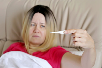 Sick woman lies in a bed with digital thermometer in hand, cold and flu season, selective focus. Concept of illness, fever, high body temperature