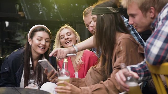 Young people using app watching photos on smartphone to and have good time in gazebo at outdoors, they laugh, joke and drink low alcohol