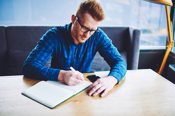 Concentrated male student in optical spectacles for provide eyes protection writing answers for school test using textbook for education on publicity area, serious hipster guy learning indoors