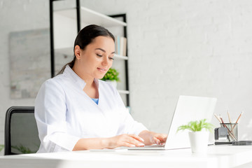 smiling dermatologist sitting at table and using laptop in clinic
