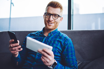 Portrait of cheerful man in eyewear enjoying leisure time indoors while sitting with modern gadgets...