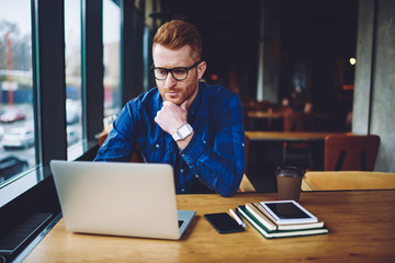 Pondering hipster guy watching important webinar for certification training in programming, serious man working remotely in coffee shop and reading received email for colleague connected to wifi