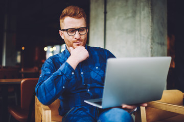 Puzzled skilled hipster guy making payment online on online web store while connected to wifi on modern netbook indoors, serious pensive man reading news about innovation in technology world