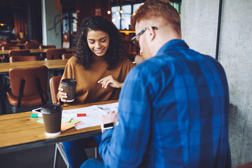 Positive multicultural male and female designers working togetherness on new architect sketches during coffee time in cafeteria, happy successful man and woman sitting at table and analyzing process