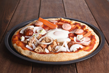pizzza with egg, onion, tuna fish, carrot and frankfurter in a backing tin, on a wooden table, freshly baked