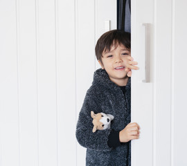 Cute kid wearing fluffy pajamas hugging dog toy playing hide and seek  in wardrobe, little boy looking out while holding the white door, Child with happy face having fun playing  with closet