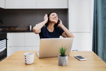 Attractive and happy middle age female freelancer is working at her home. She is using headphones, listening to music and singing. Modern kitchen in background. Freelancing job concept.