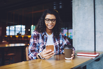 Happy african female blogger enjoying time for communication with followers from social networks using mobile application and free wifi internet in coffee shop, positive hipster girl chatting