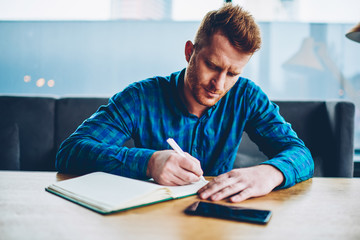 Skilled red haired student writing down homework in copybook studying at wooden table in coworking space.Pensive young man dressed in casual shirt noting checklist in notepad during free time