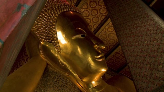 A closeup, low angle shot of the giant golden reclining Buddha's face with its head resting on its curled arm inside the Wat Pho temple in Thailand.