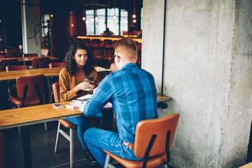 Cute african american young woman with curly hair holding coffee to go in hands and listening male architector sitting in university and working together on common project.Two students talking