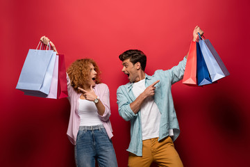 beautiful surprised couple pointing at shopping bags, isolated on red