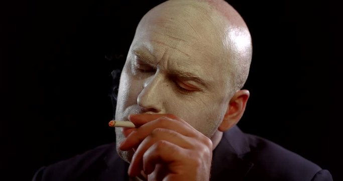 strange man with white makeup on face is lighting a cigarette and smoking, looking thoughtfully