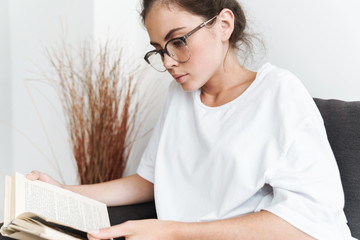 Image of serious young woman reading book while sitting on sofa