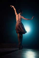 Plakat Ballerina is practicing her moves on dark stage. Young girl dancing in beige dress, spinning around and smiling. Gracefulness and tenderness in every movement.