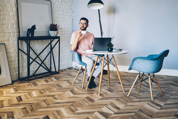 Pondering hipster guy enjoying coffee break during working remotely on laptop device in home interior with stylish design, contemplative male copywriter thinking about idea for new article on web page