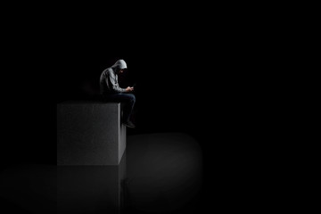  a lonely man with smartphone sits on a concrete cube in the dark. reflection on the floor. - obrazy, fototapety, plakaty