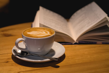 Ceramic cup of cappuccino in coffee shop with pattern on a wooden table with an open book. Latte art. Morning drink. Caffeine.