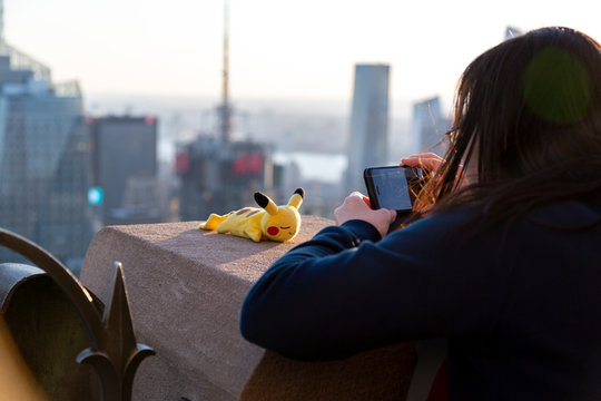 Asian girl taking a picture of Pokemon character Pikachu toy with New York City Manhattan panorama.