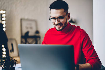 Positive young man in optical eyewear laughing while searching information on website on modern laptop computer using wireless internet.Cheerful hipster guy working remotely at netbook in apartment