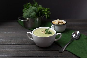 Vegetarian cream of broccoli, potato, onion, garlic, sesame and olive oil, and coconut milk soup. served in a white ceramic plate with a small pan on a dark brown wooden board using green textile