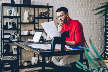 Concentrated hipster student learning information from text papers sitting at desktop in modern studio.Pensive male graphic designer looking at blank sketch and thinking on creative ideas