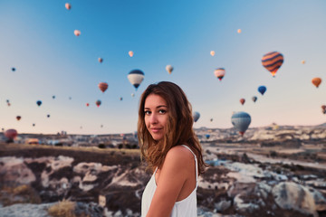 A tourist girl wearing white sweater on a mountain top enjoying wonderful view of balloons in Cappadocia