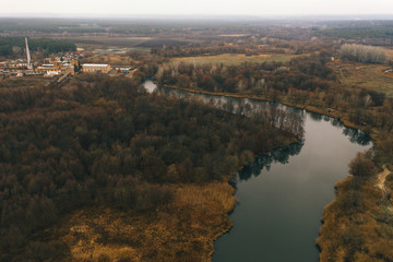 Autumn landscape view of the river, forest and factory. A curved riverbed with banks, overgrown with trees and with a plant in the distance.