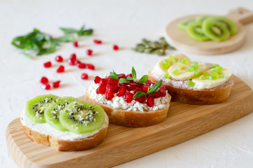 Open vegetarian sandwiches with vegetables, fruits and cream cheese on a wooden board on a white background. Kiwi and pomegranate in the background. Healthy breakfast concept. Copy space.