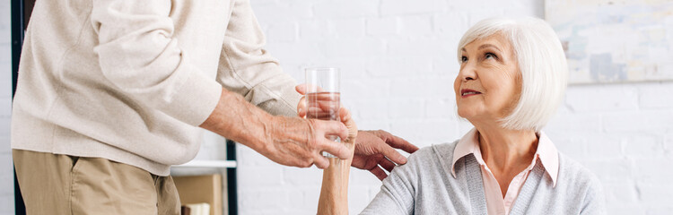 panoramic shot of husband giving glass of water to wife in apartment