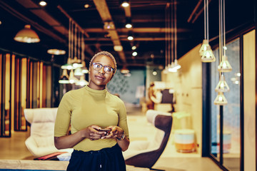 Portrait of african american young woman in eyeglasses looking at camera while chatting online on modern smartphone device standing in office interior.Dark skinned female proud ceo with cellular