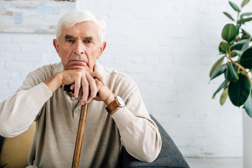 senior man looking at camera and holding wooden cane in apartment