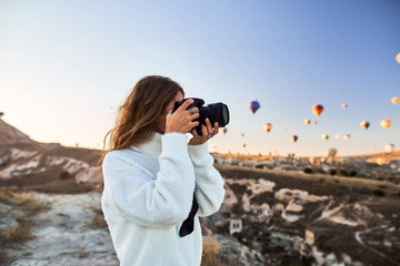 Travel photographer wearing white sweater taking a picture of balloons in Cappadocia. Travel photographer in Turkey - obrazy, fototapety, plakaty