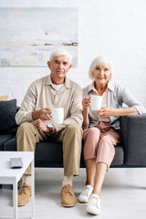 smiling husband and wife holding cups and sitting on sofa in apartment