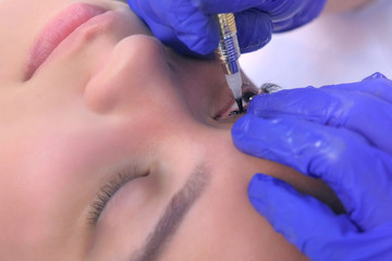 Portrait of woman on permanent eyeliner makeup with tattoo machine in beauty salon, face closeup. Cosmetologist making beauty procedure for young woman in clinic, hands closeup.