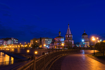 View of the Christian church through the lines from passing cars at night against a blue sky surrounded by trees, lanterns of the night city and next to the Angara embankment and its fence.