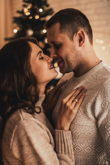 Lovely couple in sweaters hugging and celebrating new year in front of Christmas tree in decorated interior.