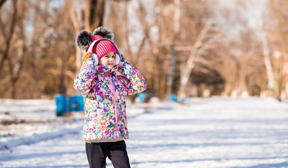 Winter portrait of smiling little girl in pink hat with bubo walk in snow park and listen to music in headphones