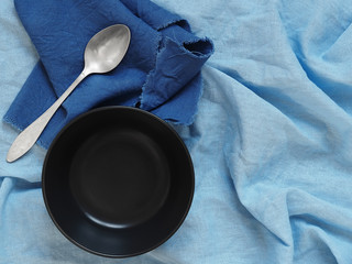 An empty black soup cup on a blue linen tablecloth, next to a vintage spoon on a dark blue napkin. Flat lay. Natural lighting. Copy space. Still life