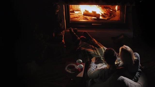 A young couple admires the fire in the fireplace, sitting next to them and drinking tea.